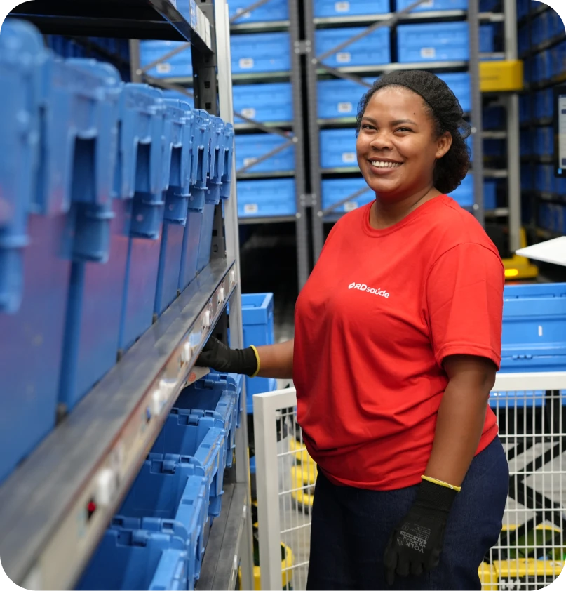 À direita, foto de uma mulher, com touca descartável e luvas, sorri e segura uma embalagem na abertura de uma caixa. Ela é negra, com cabelos cacheados presos e usa camiseta vermelha com o logotipo da R D Saúde em branco, na altura do peito esquerdo. Em frente a ela e ao fundo, diversas prateleiras metálicas e com caixas azuis empilhadas.
                    